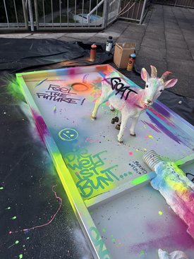 Minigolfbahn mit buntem Graffiti, darauf eine weiße Ziegenskulptur mit Schriftzug und bunte Farbspritzer