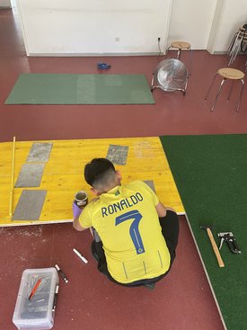 Junge mit gelbem Trikot mit Aufdruck 'RONALDO 7' sitzt auf dem Boden und arbeitet an einer gelben Holzplatte, umgeben von Werkzeugen und zwei Hockern in einem Raum mit rotem Boden.