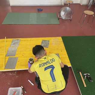 Junge mit gelbem Trikot mit Aufdruck 'RONALDO 7' sitzt auf dem Boden und arbeitet an einer gelben Holzplatte, umgeben von Werkzeugen und zwei Hockern in einem Raum mit rotem Boden.