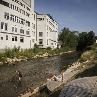 Foto: Blick auf die terassenförmig angelegten Sitz- und Liegemöglichkeiten an der Fils. Auf der anderen Seite der Fils ist ein großes Gebaüde zu sehen. Ein Kind Spielt im Wasser, eine Frau, vermutlich die Mutter, schaut vom Ufer aus zu.