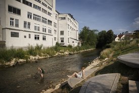 Foto: Blick auf die terassenförmig angelegten Sitz- und Liegemöglichkeiten an der Fils. Auf der anderen Seite der Fils ist ein großes Gebaüde zu sehen. Ein Kind Spielt im Wasser, eine Frau, vermutlich die Mutter, schaut vom Ufer aus zu.
