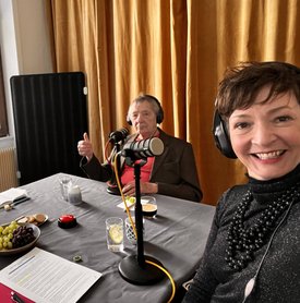 Gast Peter Grohmann und Moderatorin Kimsy von Reischach während der Aufnahme des Podcast. Beide sitzen an einem Tisch, tragen Kopfhörer und haben ein Tischmikrofon vor sich stehen. 