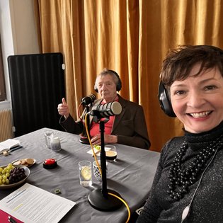 Gast Peter Grohmann und Moderatorin Kimsy von Reischach während der Aufnahme des Podcast. Beide sitzen an einem Tisch, tragen Kopfhörer und haben ein Tischmikrofon vor sich stehen.