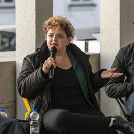 Sina Reisch auf der Terrasse des Literaturmuseum der Moderne. Sina Reisch hat kurzes, lockiges Haar und trägt dunkle Klamotten. Sie spricht in ein Mikrofon.