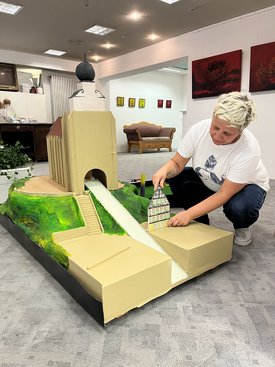 Frau kniet neben einem Minigolfmodell einer Kirche mit Turm und platziert ein kleines Modellgebäude auf dem Spielfeld