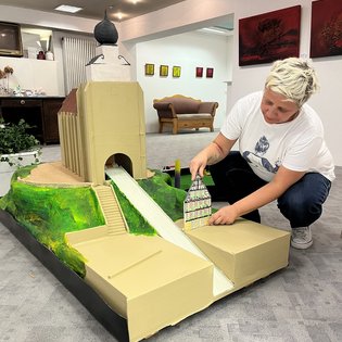 Frau kniet neben einem Minigolfmodell einer Kirche mit Turm und platziert ein kleines Modellgebäude auf dem Spielfeld