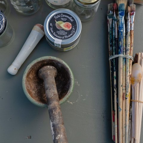 Foto von Mörser und Stößel, einer Dose mit der Aufschrift ‚Fichi Caramellati‘, mehreren Farbgläsern, benutzten Pinseln, einem Plastiklöffel und Glasbehältern