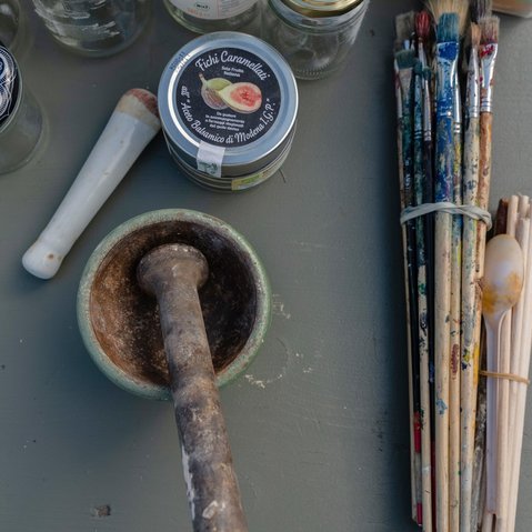 Foto von Mörser und Stößel, einer Dose mit der Aufschrift ‚Fichi Caramellati‘, mehreren Farbgläsern, benutzten Pinseln, einem Plastiklöffel und Glasbehältern