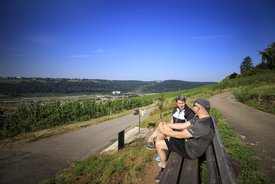 Zwei Personen - möglicherweise ien Paar - sitzen auf einer Bank und unterhalten sich. Im Hintergrund sind Weinberge und ein Ort zu sehen.