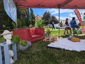 Auf einer Wiese unter einem Pavillion sind verschiedene Möbelstücke, Kunstwerke und Dekoartikel wie ein Sofa, eine Holzkiste als Tisch, eine Decke mit Kissen, Büsten und Pflanzen angeordnet.