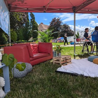 Auf einer Wiese unter einem Pavillion sind verschiedene Möbelstücke, Kunstwerke und Dekoartikel wie ein Sofa, eine Holzkiste als Tisch, eine Decke mit Kissen, Büsten und Pflanzen angeordnet.