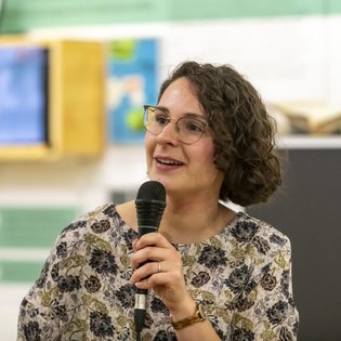 Lea Wegner hält ein Mikrofon in der Hand. Sie hat halblanges braunes, lockiges Haar, trägt eine Brille und eine Bluse mit Blumenmuster.