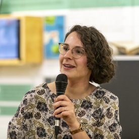 Lea Wegner hält ein Mikrofon in der Hand. Sie hat halblanges braunes, lockiges Haar, trägt eine Brille und eine Bluse mit Blumenmuster.