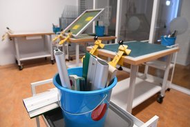 Ein heller Arbeitsraum mit mehreren Tischen, auf denen Werkzeuge und Vorrichtungen für den Siebdruck vorbereitet sind. Im Vordergrund ein blauer Eimer mit Werkzeugen wie Rakeln und Aluminiumprofilen. Die Tische sind mit Klammern ausgestattet, die Siebe halten, und im Hintergrund sind weitere Materialien und Geräte ordentlich angeordnet