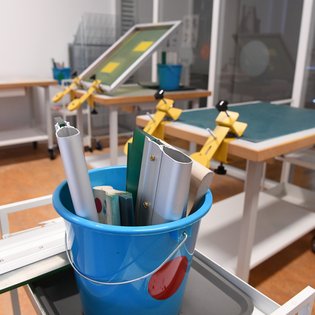 Ein heller Arbeitsraum mit mehreren Tischen, auf denen Werkzeuge und Vorrichtungen für den Siebdruck vorbereitet sind. Im Vordergrund ein blauer Eimer mit Werkzeugen wie Rakeln und Aluminiumprofilen. Die Tische sind mit Klammern ausgestattet, die Siebe halten, und im Hintergrund sind weitere Materialien und Geräte ordentlich angeordnet
