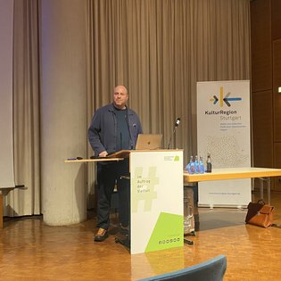 Impulsgeber Jacob Sylvester Bilabel steht an einem Rednerpult in einem Veranstaltungssaal, vor einer Projektionswand und einem Banner der KulturRegion Stuttgart, mit Laptop und Mikrofon.