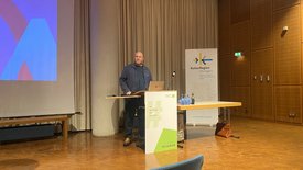 Impulsgeber Jacob Sylvester Bilabel steht an einem Rednerpult in einem Veranstaltungssaal, vor einer Projektionswand und einem Banner der KulturRegion Stuttgart, mit Laptop und Mikrofon.