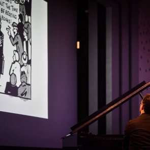 Pianist Itay Dvori sitzt an einem Fügel. Während er spielt schaut er auf eine Leinwand auf die ein Comic projiziert wird.