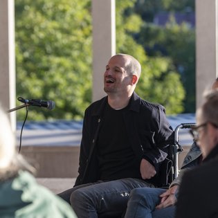 Michael Turinsky auf der Terrasse des Literaturmuseum der Moderne. Turinsky trägt eine dunkle Jacke und ein dunkles Shirt. Er sitzt vor einem Mikrofon und nutzt einen Rollstuhl.