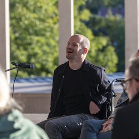 Michael Turinsky auf der Terrasse des Literaturmuseum der Moderne. Turinsky trägt eine dunkle Jacke und ein dunkles Shirt. Er sitzt vor einem Mikrofon und nutzt einen Rollstuhl.