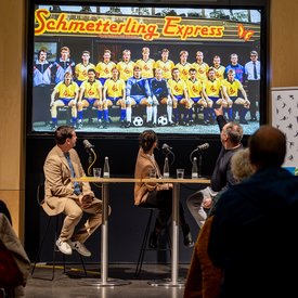 Moderator Ingmar Volkmann, Rocío Alemán und Alexander Zorniger während der Podcast Aufnahme. Alle drei sitzen auf Barhockern an Tischen und haben Mikrofone vor sich. Alle drei blicken im Moment der Aufnahme nach hinten auf eine Videoleinwand, auf der das Foto einer Fußballmannschaft zu sehen ist. Alexander Zorniger deutet auf eine Person auf dem Bild.