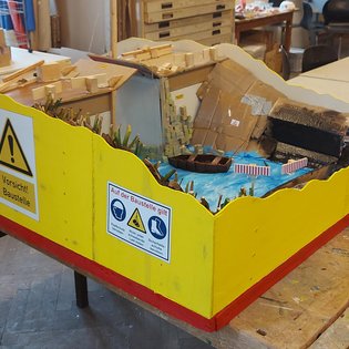 Modell einer Minigolfbahn mit gelb-roter Umrandung und Wasserhindernis auf Werkbank in Werkstatt.