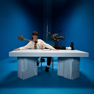 Person mit weißem Hemd und Krawatte sitzt an einem Tisch mit zwei Stapeln Papier als Tischbeine vor blauem Hintergrund, eine Filmkamera ist hinter der Person sichtbar