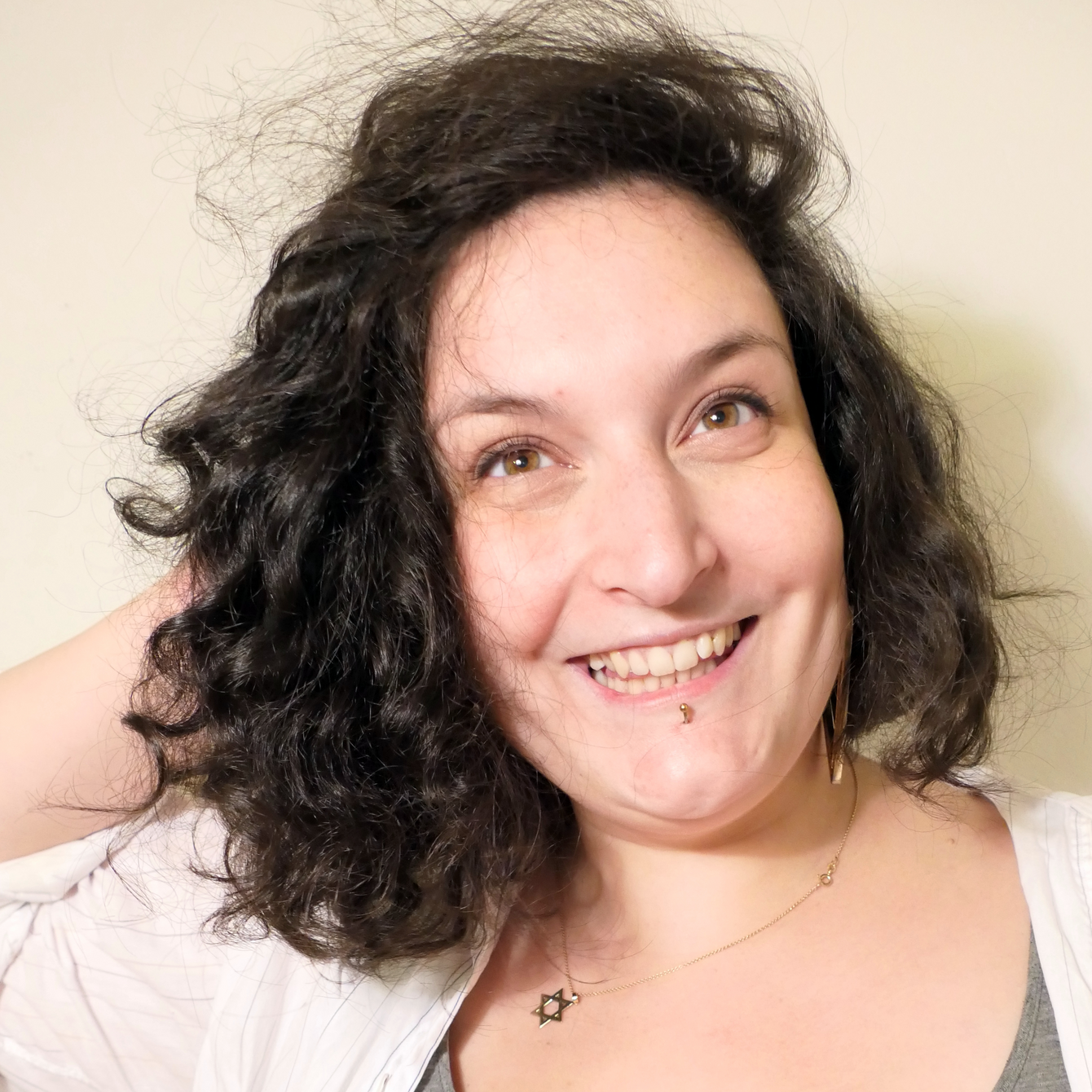 Portraitbild von Debora Antmann. Debora Antmann hat halblanges, dunkles, lockiges Haar und braune Augen. Auf dem Bild trägt sie eine weiße Bluse über einem grauen Shirt, eine feine goldene Kette mit einem Davisstern-Anhänger und ein Piercing an der Lippe. Sie lacht fröhlich und hat den Blick über die Kamera hinweg gerichtet