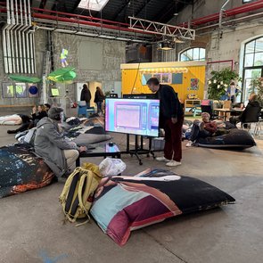Mehrere Personen sitzen und liegen auf großen Sitzkissen in einer Industriehalle mit sichtbaren Rohren und einem gelben Bauwagen im Hintergrund