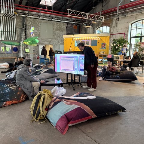 Mehrere Personen sitzen und liegen auf großen Sitzkissen in einer Industriehalle mit sichtbaren Rohren und einem gelben Bauwagen im Hintergrund