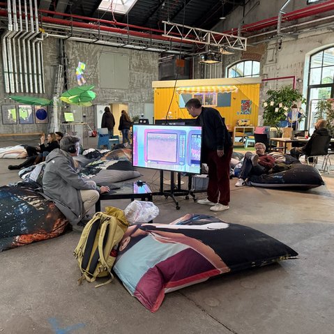 Mehrere Personen sitzen und liegen auf großen Sitzkissen in einer Industriehalle mit sichtbaren Rohren und einem gelben Bauwagen im Hintergrund
