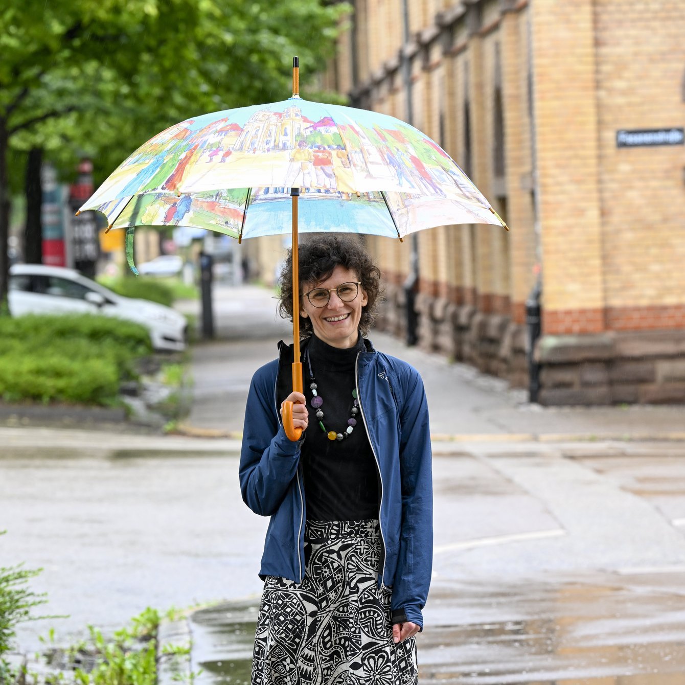 Lucia Leidenfrost an einem regnerischen Tag mit buntem Schirm in einer städtischen Umgebung.