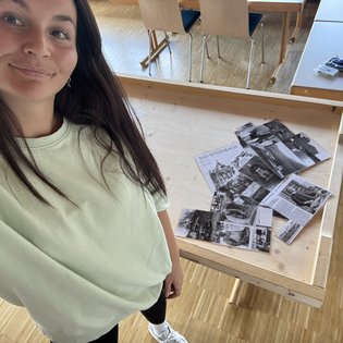 Frau in hellem T-Shirt macht Selfie vor einem Holztisch mit mehreren schwarz-weißen Fotos und einem Blatt mit der Überschrift 'BAD DITZENBACH'