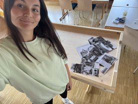 Frau in hellem T-Shirt macht Selfie vor einem Holztisch mit mehreren schwarz-weißen Fotos und einem Blatt mit der Überschrift 'BAD DITZENBACH'