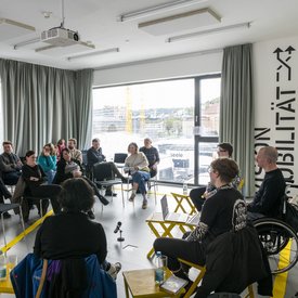 Im Vordergrund Michael Turinsky mit seinen Gäst*innen. Turinsky nutzt einen Rollstuhl, alle anderen sitzen auf gelben Stühlen. Im Hintergrund sitzt das Publikum. Eine große Panoramascheibe bietet einen Blick auf die Stuttgart 21-Baustelle.