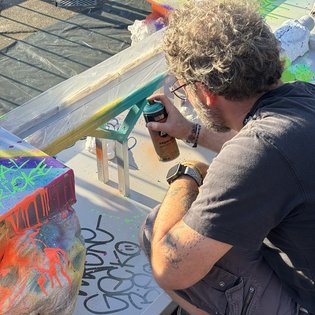 Mann mit lockigem Haar und Brille sprüht mit Farbdose auf eine Minigolfbahn mit bunten Farbspritzern