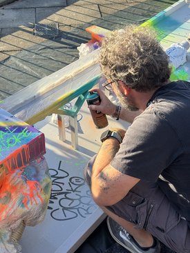 Mann mit lockigem Haar und Brille sprüht mit Farbdose auf eine Minigolfbahn mit bunten Farbspritzern