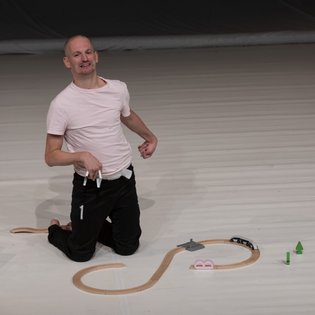 Michael Turinsky auf der Bühne in einer Tanzpose auf den Knien. Er trägt ein helles Shirt und eine schwarze Hose. Vor ihm eine halb zusammengebaute Spielzeug-Holzeisenbahn.