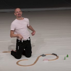Michael Turinsky auf der Bühne in einer Tanzpose auf den Knien. Er trägt ein helles Shirt und eine schwarze Hose. Vor ihm eine halb zusammengebaute Spielzeug-Holzeisenbahn.