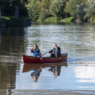 Zwei weiblich gelesene Personen in einem Kanu auf einem Gewässer. Beide tragen Schwimmwesten und halten je ein Paddel in den Händen.
