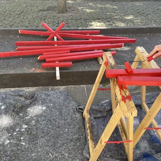Mehrere rot lackierte Holzlatten liegen auf dem Boden und einem Holzgestell, eine Hand hält einen Pinsel und lackiert eine weitere Latte rot
