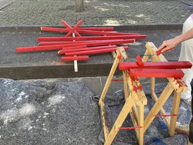 Mehrere rot lackierte Holzlatten liegen auf dem Boden und einem Holzgestell, eine Hand hält einen Pinsel und lackiert eine weitere Latte rot