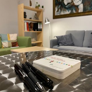 Nahaufnahme eines Metalltisches mit drei schwarzen Stiften und einem Stapel quadratischer Flyer der Artothek Filderstadt. Im Hintergrund ein gemütlicher Raum mit einem grauen Sofa, einem grünen Sessel, einem Holzregal mit Büchern und Skulpturen sowie einem großen Gemälde an der Wand. Die Atmosphäre wirkt einladend und kreativ.