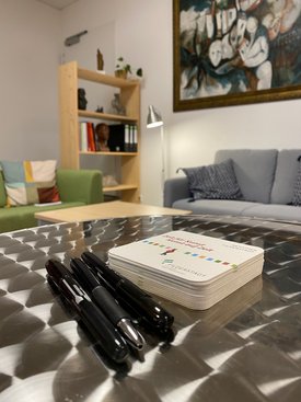 Nahaufnahme eines Metalltisches mit drei schwarzen Stiften und einem Stapel quadratischer Flyer der Artothek Filderstadt. Im Hintergrund ein gemütlicher Raum mit einem grauen Sofa, einem grünen Sessel, einem Holzregal mit Büchern und Skulpturen sowie einem großen Gemälde an der Wand. Die Atmosphäre wirkt einladend und kreativ.