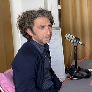 Dr. Robert Ogman während der Podcast-Aufnahme. Dr. Ogman hat dunkles, lockiges Haar und er trägt ein dunkles Hemd mit einer dunklen Weste. Vor ihm steht ein Mikrofon auf einem Tisch.