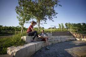 Zwei Personen, möglicherweise ein Paar - sitzen auf halbrund angeordneten Steinen und unterhaten sich. Im Hintergrund ist ein eg durch die Weinberge zu sehen.