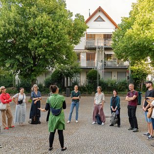 ca 15 Personen stehen auf einem Platz im Halbkreis, die Frau im grünen Hemd erklärt etwas vor ihnen stehend