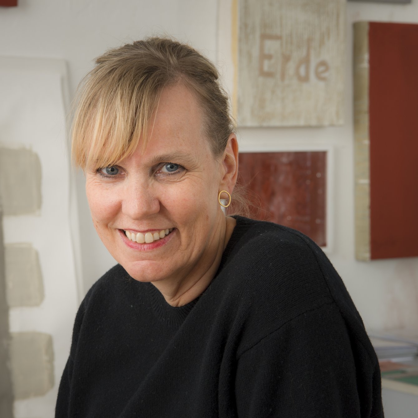 Auf dem Bild ist die bildende Künstlerin und Gründerin des Projektraums kunst [ ] klima Barbara Karsch-Chaïeb zu sehen. Ihre blonden Haare sind bis auf den Pony streng nach hinten zurückgekämmt. Sie trägt ein schwarzes Oberteil und lächelt in die Kamera. Im Hintergrund sind einige ihrer Kunstwerke an der Wand zu erkennen, alle in erdigen Brauntönen gehalten.