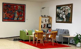 Ein freundlich gestalteter Sitzbereich in der Artothek Filderstadt. Auf einem roten Teppich stehen vier Korbstühle um einen kleinen Holztisch, flankiert von einem grünen Sessel und einem grauen Sofa mit bunten Kissen. Dahinter befindet sich ein Holzregal mit Büchern, Skulpturen und Pflanzen. Zwei große, farbintensive Gemälde hängen an den weißen Wänden. Die Atmosphäre wirkt offen, kreativ und einladend.