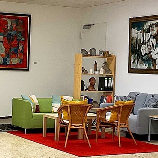 Ein freundlich gestalteter Sitzbereich in der Artothek Filderstadt. Auf einem roten Teppich stehen vier Korbstühle um einen kleinen Holztisch, flankiert von einem grünen Sessel und einem grauen Sofa mit bunten Kissen. Dahinter befindet sich ein Holzregal mit Büchern, Skulpturen und Pflanzen. Zwei große, farbintensive Gemälde hängen an den weißen Wänden. Die Atmosphäre wirkt offen, kreativ und einladend.